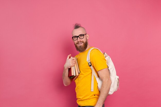 Estudiante adulto alegre chico de ropa casual con barba y mochila sosteniendo libros aislados en rosa