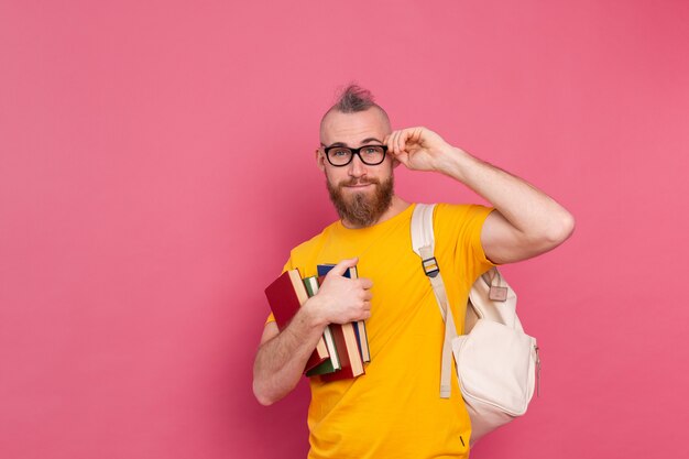 Estudiante adulto alegre chico de ropa casual con barba y mochila sosteniendo libros aislados en rosa