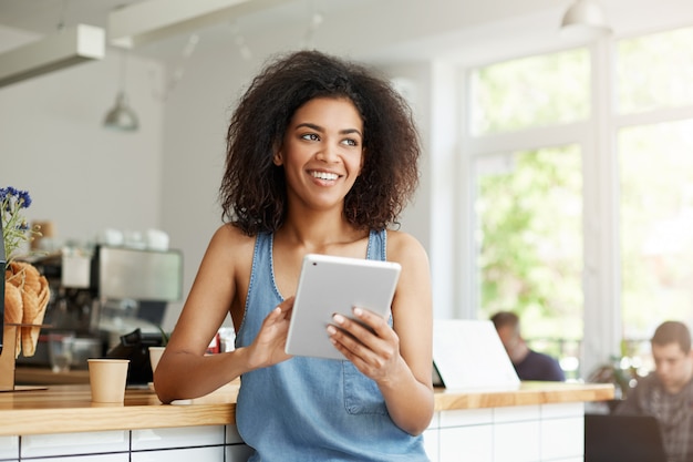 Estudiante africano hermoso alegre que descansa en café que sonríe mirando en tableta que se sostiene lateral.