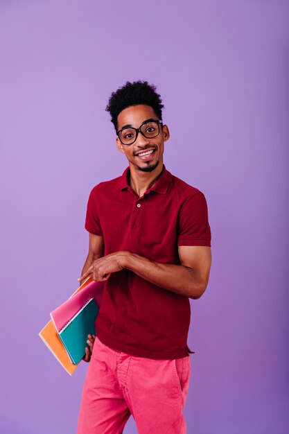 Estudiante africano de ojos oscuros posando con sus libros. chico inteligente en grandes vasos de moda aislados.