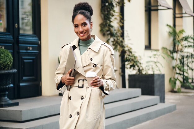 Una estudiante afroamericana bastante informal con una elegante gabardina con portátil y café para ir mirando alegremente a la cámara en la calle de la ciudad