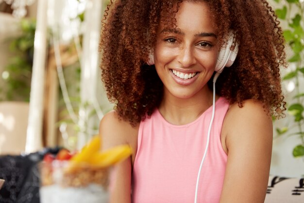 Estudiante afroamericana escucha canciones de la lista de reproducción, disfruta de un sonido perfecto en auriculares modernos, tiene una expresión alegre, se sienta contra el interior del café. Personas, ocio, concepto de entretenimiento.