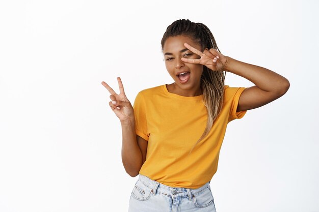 Una estudiante afroamericana positiva muestra un saludo de paz y sonríe guiños a la cámara feliz posando sobre fondo blanco