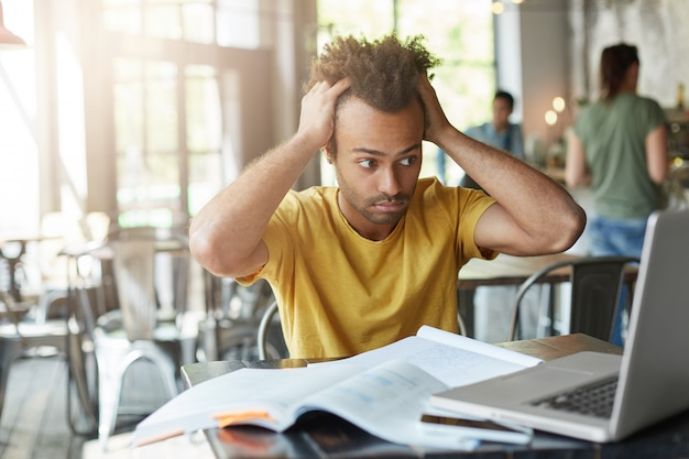 Estudiante afroamericano internacional sintiéndose estresado, manteniendo las manos en la cabeza, mirando la pantalla del portátil con frustración y desesperación