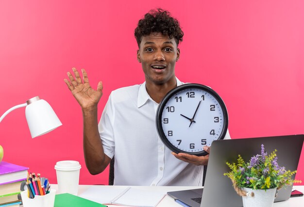 Estudiante afroamericano joven sorprendido sentado en el escritorio con herramientas escolares con reloj