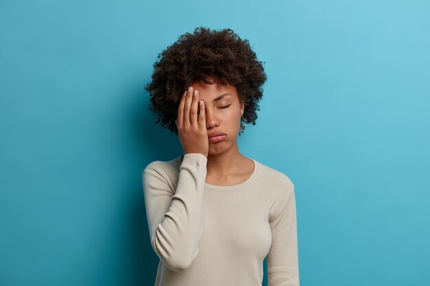 Una estudiante agotada y con exceso de trabajo hace la palma de la cara, cierra los ojos por el cansancio, pasa la noche sin dormir preparándose para los exámenes, posa infeliz en el interior, usa un jersey blanco informal, aislado en una pared azul