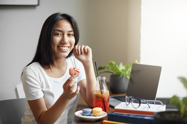 Foto gratuita estudiante asiática comiendo pastel de macarrón y bebiendo limonada mientras trabaja en su diploma con libros a su alrededor.