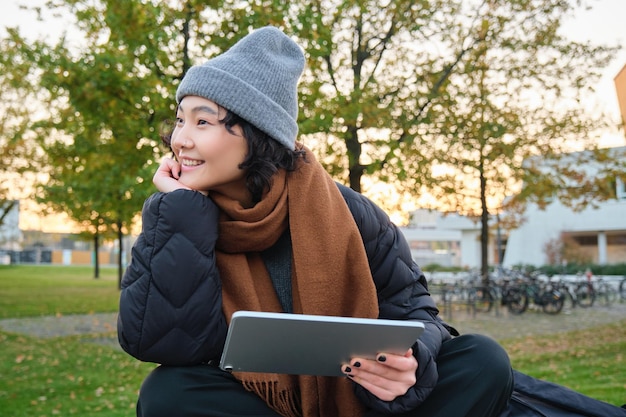 Foto gratuita una estudiante asiática sonriente se sienta en un banco en el parque sola leyendo usando la aplicación de redes sociales en la pestaña digital