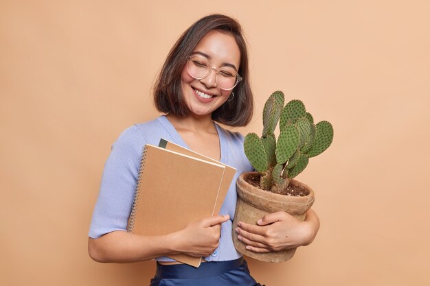 Estudiante asiático bastante alegre se siente satisfecho después de aprobar el examen lleva cuadernos y cactus en macetas usa gafas transparentes poses de puente informal contra la pared beige