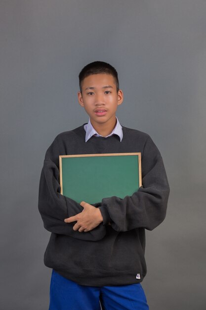 Un estudiante asiático masculino que llevaba un suéter negro abrazando el tablero verde y de pie sobre un gris.