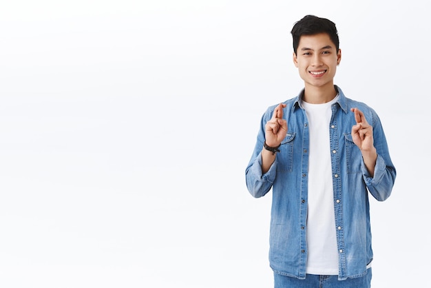 Un estudiante asiático optimista, apuesto y seguro de sí mismo, espera conseguir este trabajo buscando oportunidades de carrera, cruzar los dedos, buena suerte, sonriendo, anticipando resultados positivos, fondo blanco.