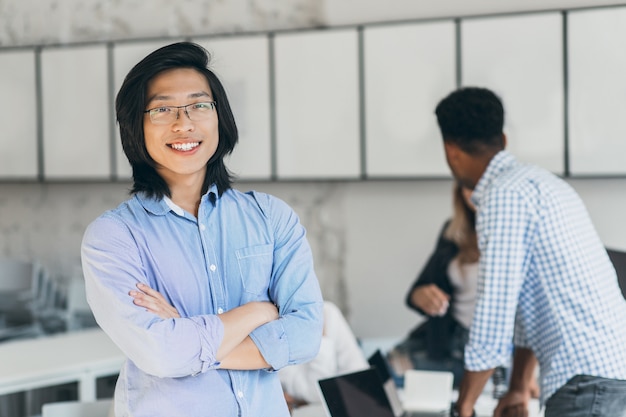 Estudiante asiático satisfecho con el pelo largo de pie en pose de confianza en la sala de conferencias. Retrato de espalda de chico africano hablando con compañeros de la universidad mientras el exitoso joven chino posando con una sonrisa.