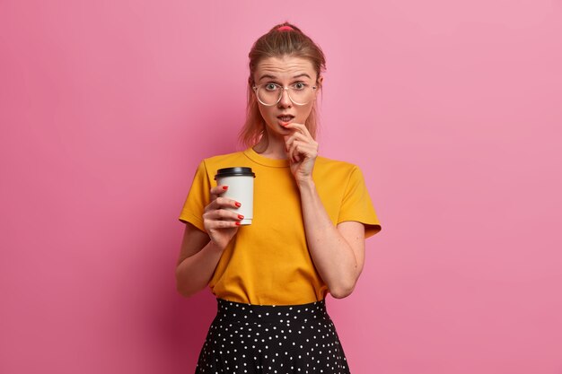 Estudiante aturdida toma café durante el recreo, tiene expresión de asombro, mira con incredulidad, sale durante el tiempo libre, usa lentes transparentes, ropa de moda