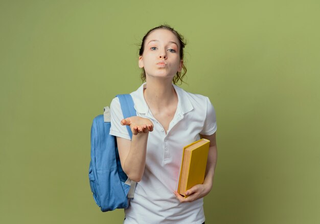 Estudiante bastante joven confidente que lleva el bolso trasero que sostiene el libro y que envía el beso del golpe a la cámara aislada en fondo verde con el espacio de la copia