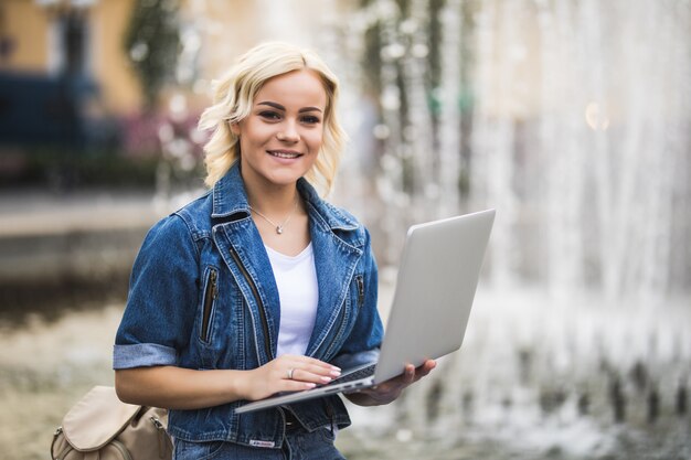 Estudiante bastante rubia chica trabaja en su computadora portátil cerca de la fuente en la ciudad en el día