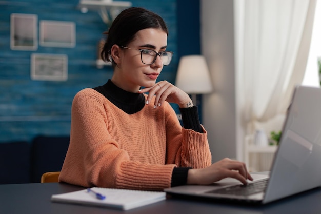 Estudiante confiado que estudia a distancia mirando la pantalla del portátil. Creador de contenido con gafas escribiendo casualmente sentado en el escritorio de casa. Chica universitaria navegando por internet. Freelancer trabajando desde casa enviando