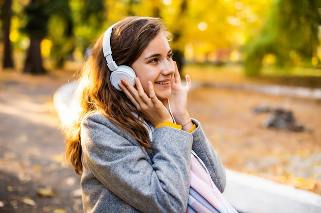 Estudiante contento joven adolescente feliz sentado al aire libre en el hermoso parque de otoño escuchando música con auriculares.
