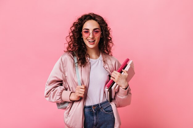 Estudiante elegante en gafas de sol posa con cuadernos sobre fondo rosa