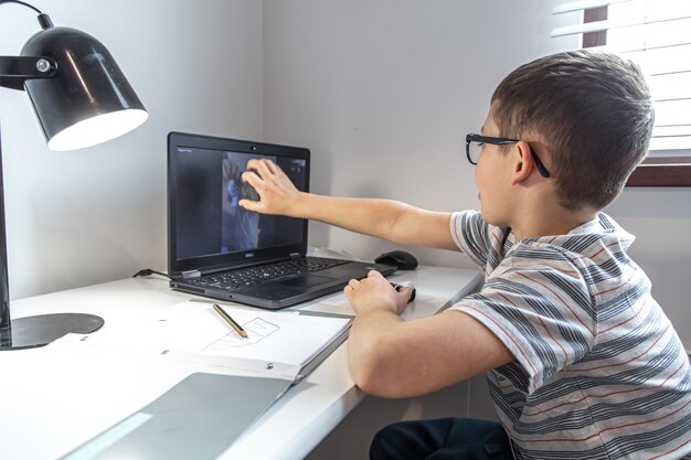 Un estudiante de escuela primaria se sienta en un escritorio frente a una computadora portátil y se comunica con un amigo a través de un enlace de video en línea, en casa.