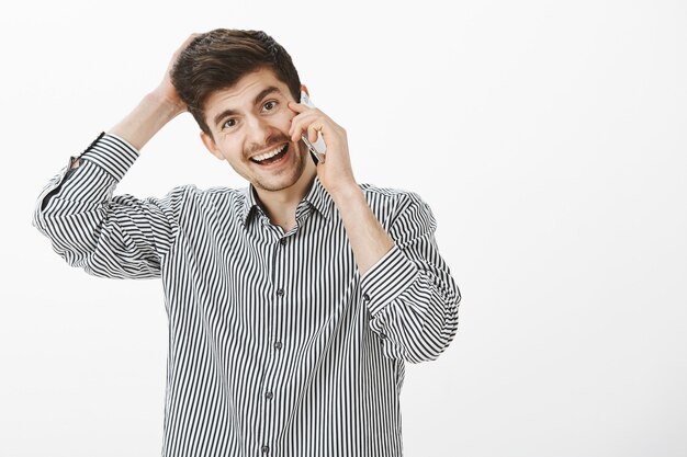 Estudiante europeo atractivo de aspecto amigable con barba y bigote, rascándose la cabeza y sonriendo ampliamente mientras habla por teléfono inteligente, olvidando algo y sintiéndose incómodo pidiendo un favor