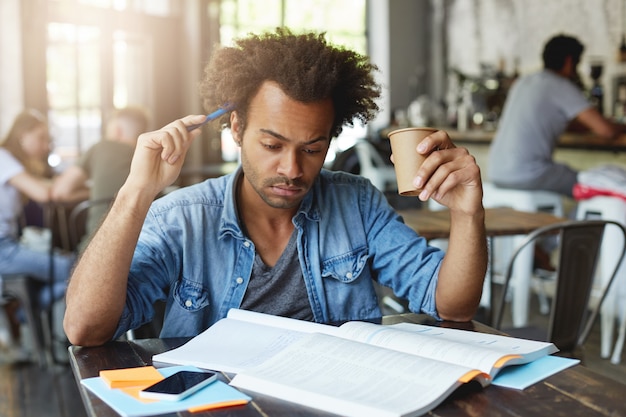 Estudiante europeo negro reflexivo concentrado con peinado afro rascándose la cabeza con un bolígrafo, bebiendo té caliente en el café, preparándose para la lección de francés en la universidad, traduciendo el artículo en un libro de texto