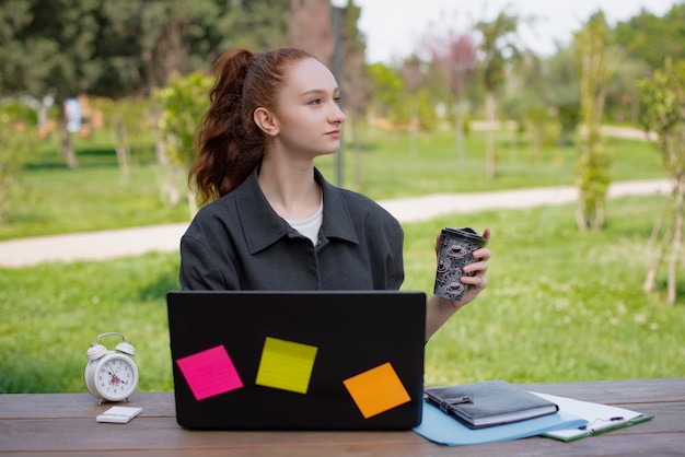 Estudiante freelance trabajando en el parque sosteniendo café bebiendo mirando hacia arriba