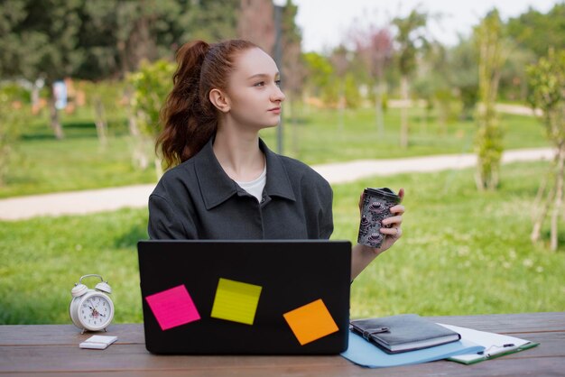 Estudiante freelance trabajando en el parque sosteniendo café bebiendo mirando hacia arriba