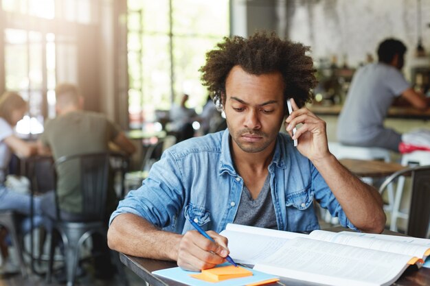 Estudiante graduado africano masculino con piel oscura y cabello exuberante trabajando en su papel de diploma escribiendo ideas clave sentado en un escritorio de madera en un café consultando a su supervisor de investigación a través de un teléfono inteligente