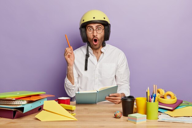 Estudiante hombre asombrado escribe algunas ideas en el cuaderno, levanta el brazo con el bolígrafo, usa casco en la cabeza, gafas, bebe café para llevar, rodeado de la papelería necesaria en el lugar de trabajo, toma notas