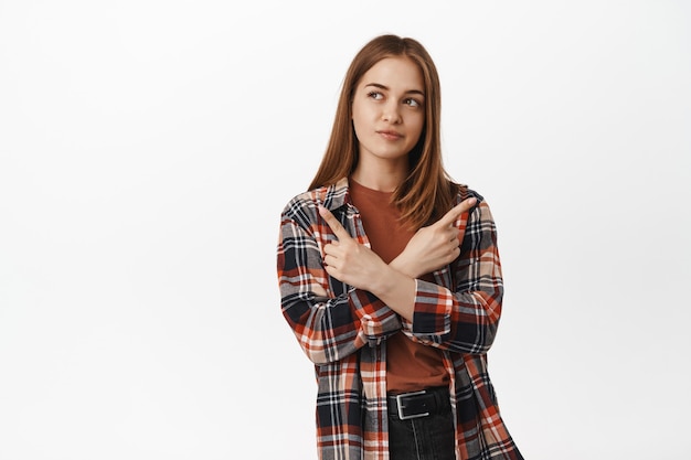 Estudiante indecisa pensando, apuntando hacia los lados y en dos direcciones, mordiéndose el labio pensativo, toma decisiones, eligiendo entre variantes, de pie contra la pared blanca