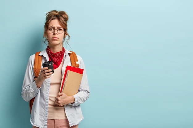 Estudiante insatisfecho con sueño posa con libros, bloc de notas y café, se siente con exceso de trabajo preparándose para el examen