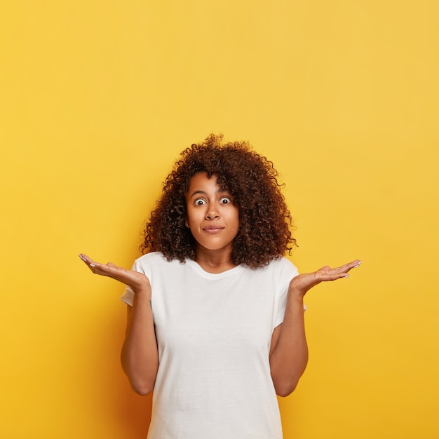 Foto gratuita una estudiante insegura y desconcertada extiende las palmas de las manos hacia los lados, mira fijamente con los ojos saltones, no tiene ni idea ni se da cuenta, tiene el pelo oscuro y rizado, usa una camiseta blanca, posa contra una pared amarilla, espacio libre hacia arriba