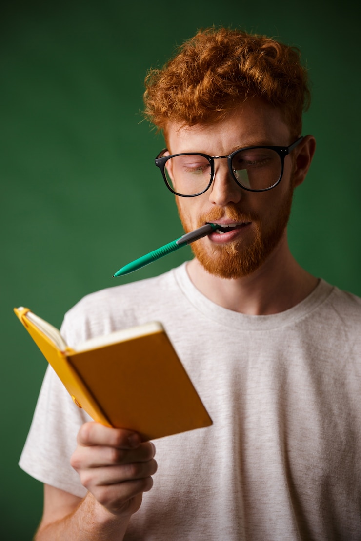 Foto gratuito estudiante inteligente de cabeza lectora con gafas con bolígrafo en la boca, sosteniendo el cuaderno