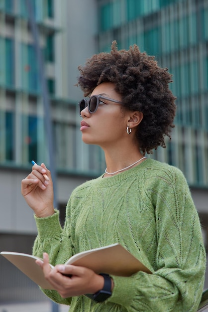 El estudiante inteligente escribe ideas en el bloc de notas para el ensayo, usa gafas de sol de moda y un jersey verde sostiene el planificador, coloca información o soportes de pensamiento al aire libre