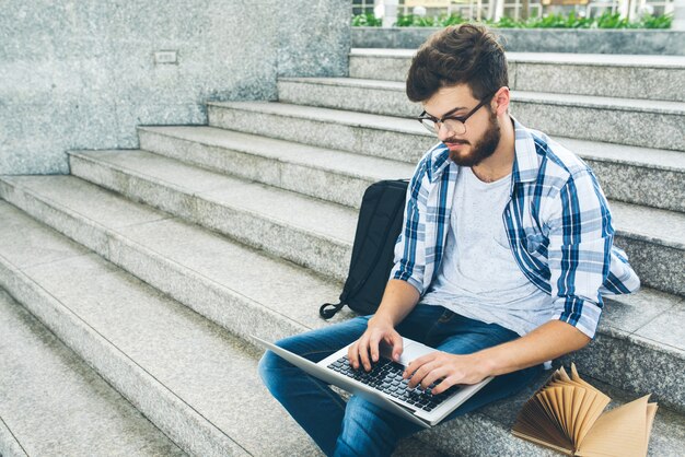 Estudiante inteligente escribiendo en el teclado de la computadora portátil descansando en las escaleras del campus al aire libre
