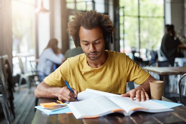 Estudiante inteligente de piel oscura que escribe algo de un libro y escucha un audiolibro en sus auriculares mientras está sentado en la cafetería durante su descanso bebiendo café para llevar y trabajando duro