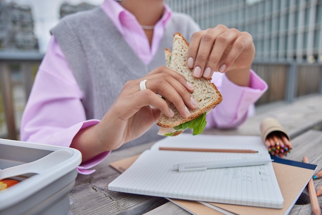 Estudiante irreconocible tiene un descanso para almorzar come posturas de sándwich en la mesa con bolígrafos abiertos y lápices de colores aprende idiomas extranjeros afuera