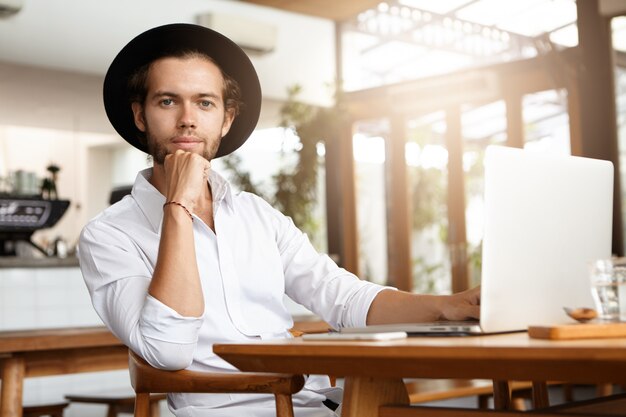 Estudiante joven atractivo alegre con sombrero negro de moda disfrutando de conexión inalámbrica gratuita, navegando por internet, usando su computadora portátil en el café