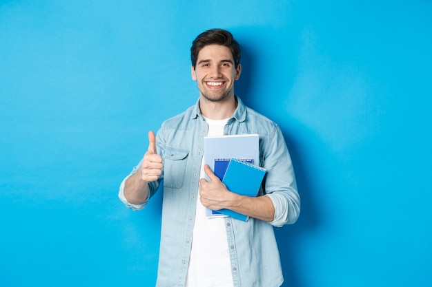 Foto gratuita estudiante joven con cuadernos, mostrando el pulgar hacia arriba en señal de aprobación, sonriendo satisfecho, fondo azul del estudio