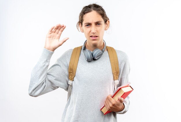 Estudiante joven disgustado con mochila con auriculares en el cuello sosteniendo libros