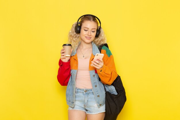 Estudiante joven en ropa moderna sosteniendo café y teléfono en amarillo
