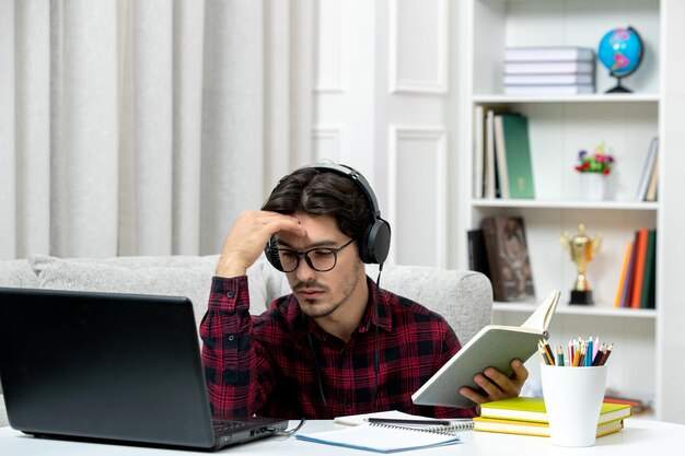 Estudiante en línea chico lindo en camisa a cuadros con gafas estudiando en la computadora cansado y confundido