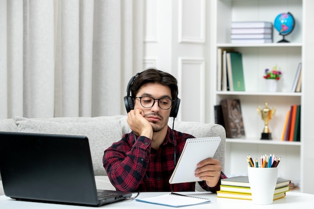 Estudiante en línea chico lindo en camisa a cuadros con gafas estudiando en la computadora cansado y leyendo