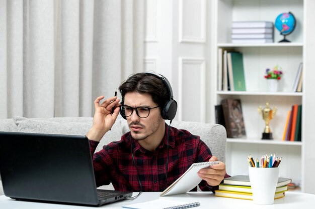 Estudiante en línea chico lindo en camisa a cuadros con gafas estudiando en la computadora confundido y concentrado
