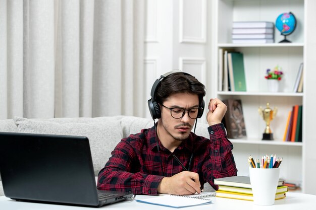 Estudiante en línea chico lindo en camisa a cuadros con gafas estudiando en la computadora confundido y escribiendo