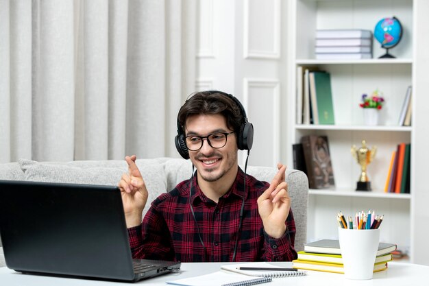 Estudiante en línea chico lindo en camisa a cuadros con gafas estudiando en la computadora cruzando los dedos