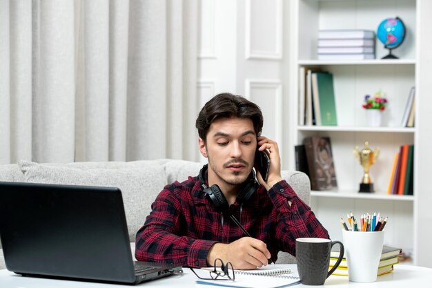 Estudiante en línea chico lindo en camisa a cuadros con gafas estudiando en la computadora escribiendo notas