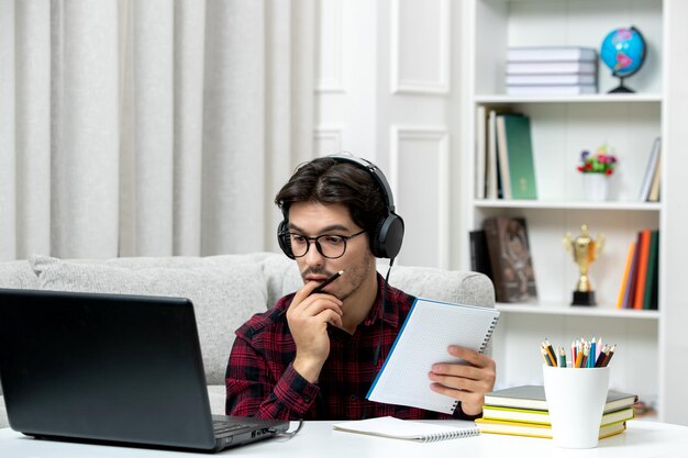 Estudiante en línea chico lindo en camisa a cuadros con gafas estudiando en la computadora escuchando