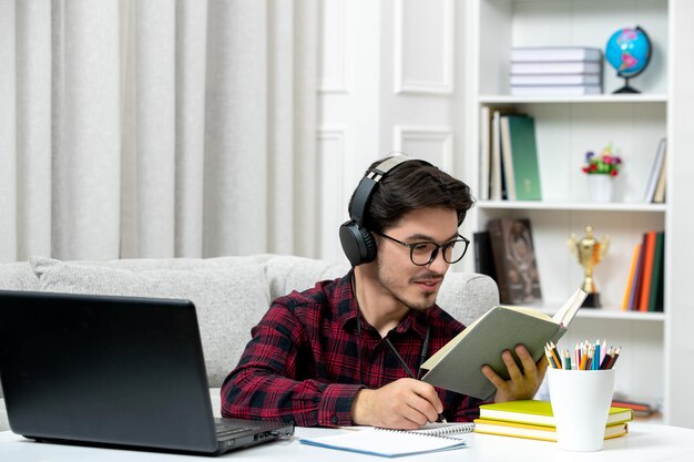 Estudiante en línea chico lindo en camisa a cuadros con gafas estudiando en la computadora leyendo un libro
