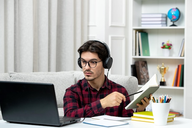 Estudiante en línea chico lindo en camisa a cuadros con gafas estudiando en la computadora leyendo un libro